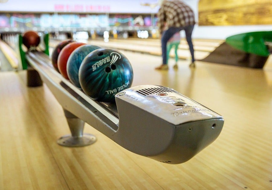 Multicolor bowing balls line up waiting to be picked while a parent helps their child set to bowl in a lane in the background