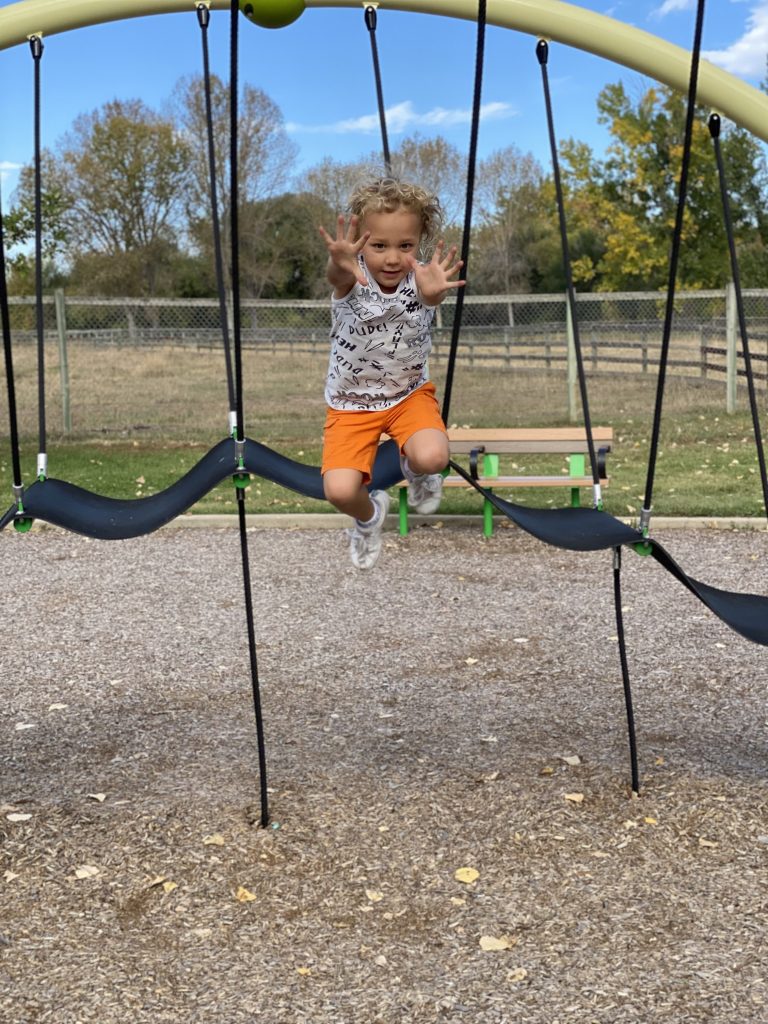 A young boy with curly hair is enthusiastically playing on a swing at a playground. He is mid-jump, with his arms extended forward and a big smile on his face.